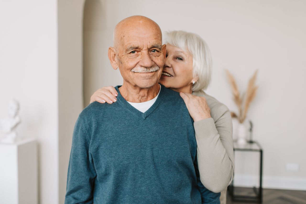 who-we-are-03 A joyful elderly couple embracing each other in a cozy indoor setting, exuding love and happiness.