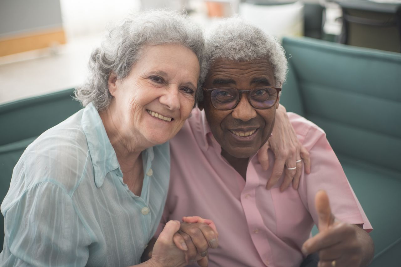 who-we-are-01 Elderly couple sharing a joyful moment indoors, sitting together on a couch with loving expressions.